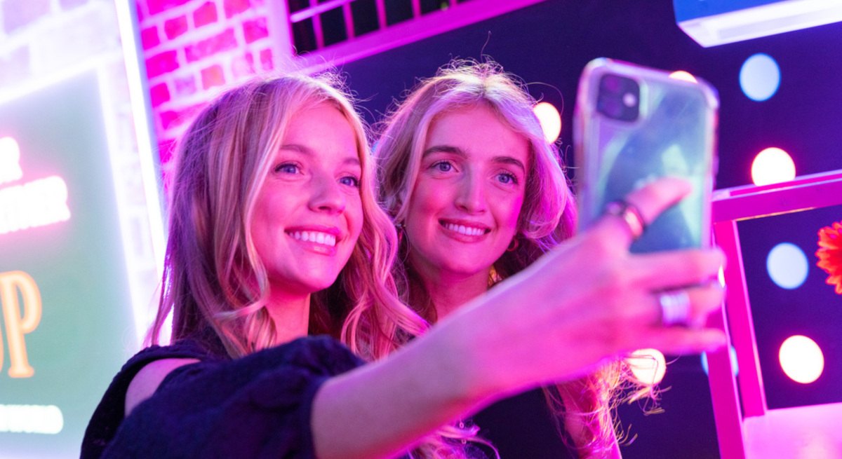 Two young women taking a selfie with a mobile phone. They are lit up with pink lighting.