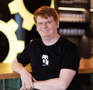 A mean with red hair and glasses, wearing a black t-shirt, leans against a counter and smiles at the camera.