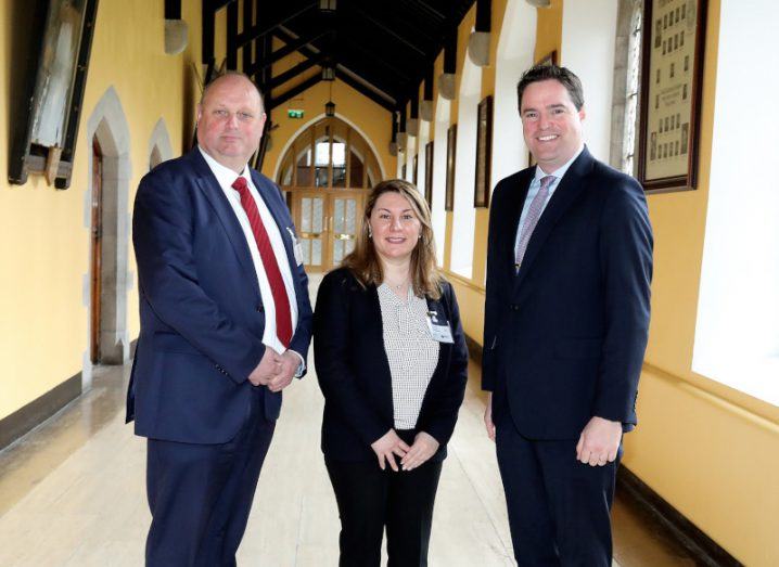 Two tall men in dark suits flank a woman with long fair hair. They are standing in the corridor of an old building with yellow walls , old portraits and large windows.