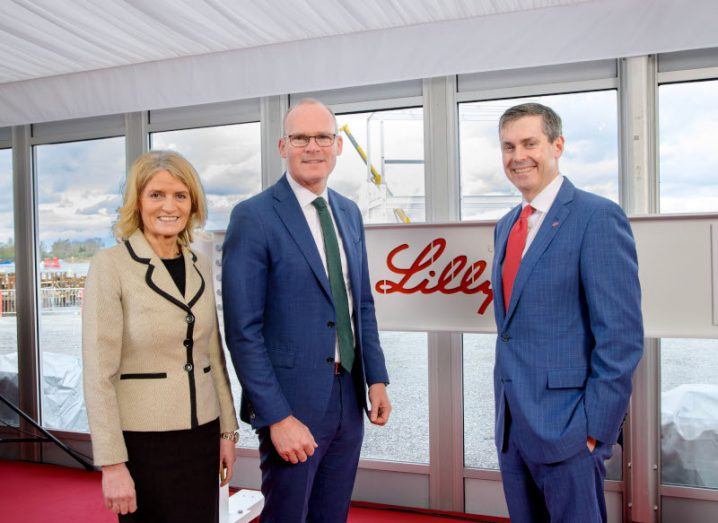 Two men and one woman standing in a room, with the Eli Lilly logo and windows behind them.