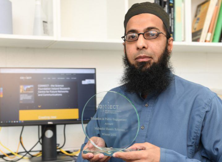 A man with glasses and a beard holding a glass award sits in an office with a computer monitor and shelves with books behind him. He is Dr Mubashir Husain Rehmani.
