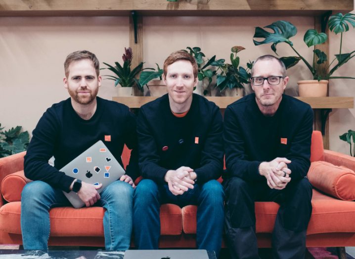 Three men in black jumpers sit on a red couch smiling at the camera, with one holding a laptop. They are the three co-founders of WhenThen.