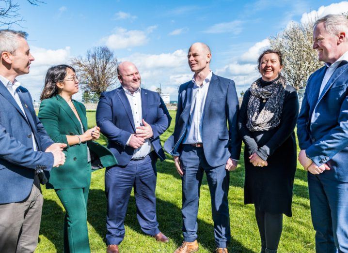 Four men and two women standing next to each other in an open grass area, smiling.