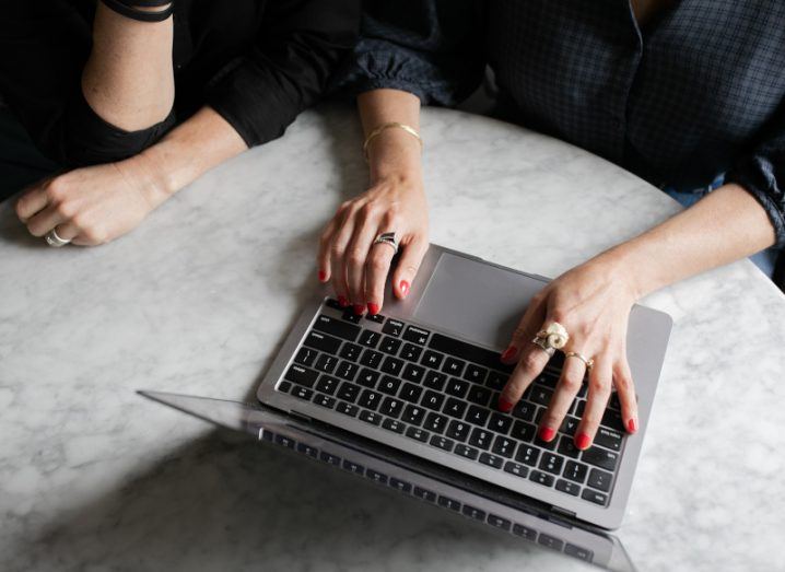 A woman's hands typing on a laptop keyboard with another woman's hands visible on the table.