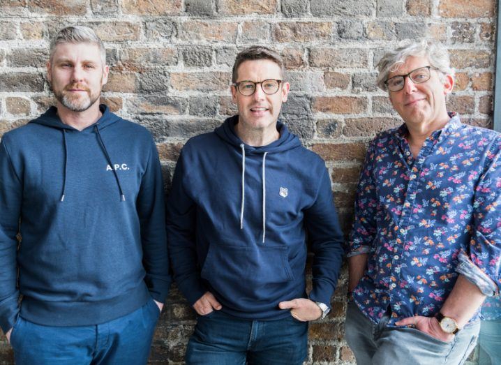 The three men who co-founded Dimply stand next to each other in front of a brick wall.