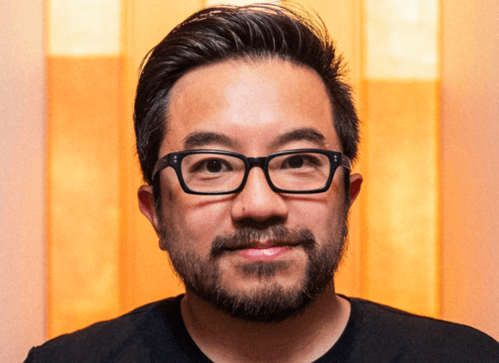 Headshot of Garry Tan wearing a black t-shirt and glasses. Yellow wall in the background.