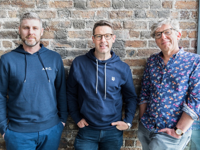 The three men who co-founded Dimply stand next to each other in front of a brick wall.