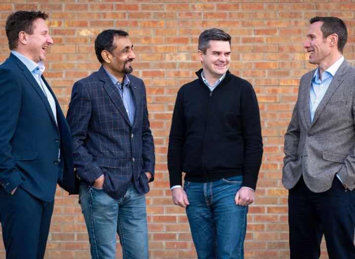 Four men stand next to each other and laugh. Brick wall in the background.