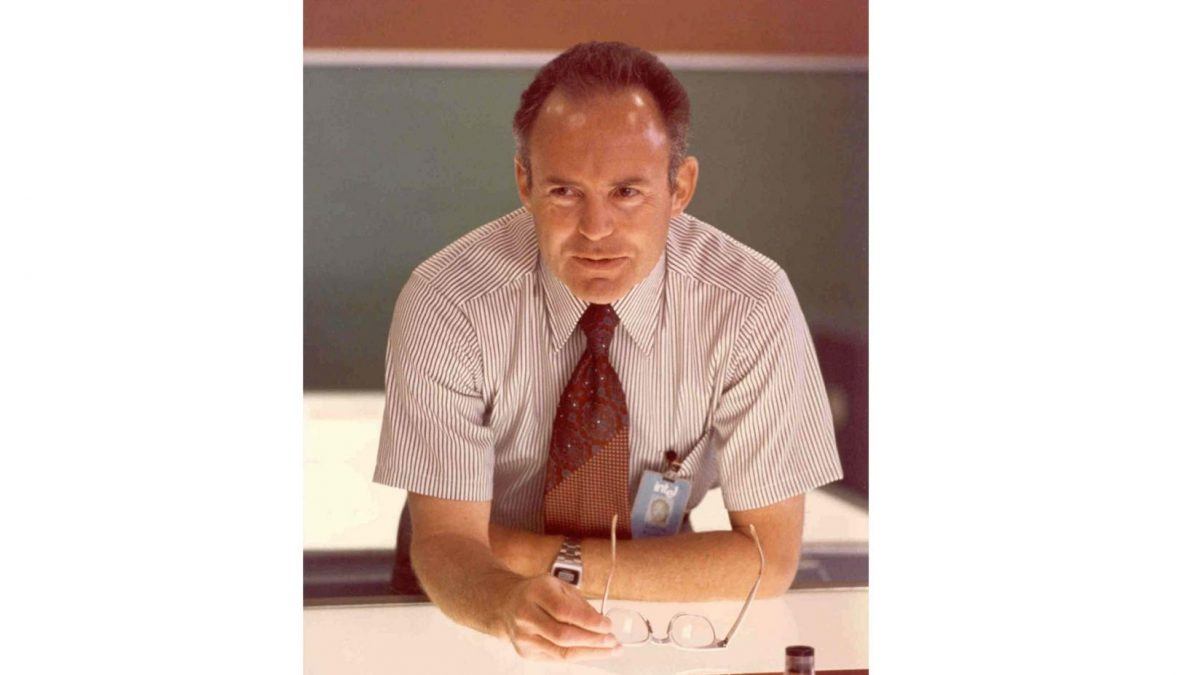 A photo of Intel co-founder Gordon Moore, leaning on the table and holding a pair of glasses in his right hand.
