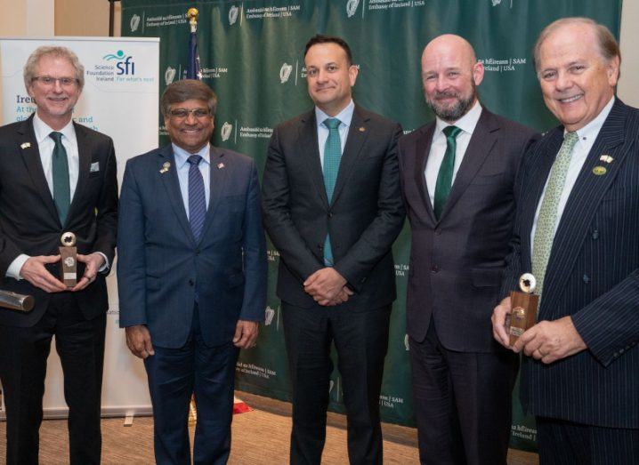 Five men stand smiling at the camera. They are all wearing suits. The SFI and Irish government banners are behind them.