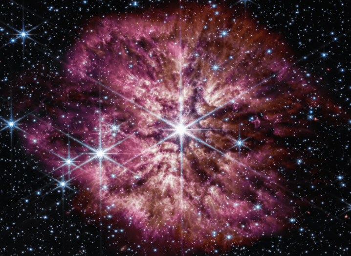 Image of stars in the universe, with a large star in the center surrounded by red and pink gases.