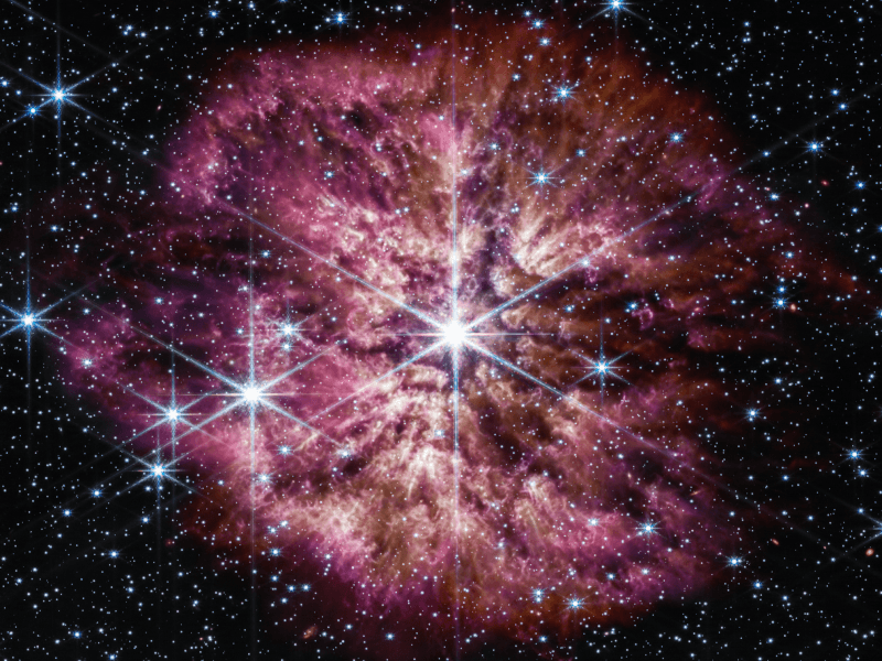 Image of stars in the universe, with a large star in the centre surrounded by red and pink gases.