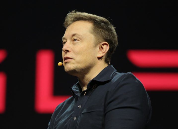 A man wearing a black shirt with a small microphone attached to his head. He is Elon Musk.