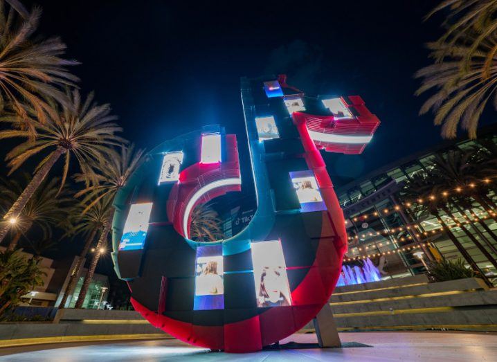 A statue of the TikTok logo, in a courtyard at night with palm trees on either side of it.