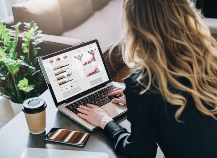 A woman typing on a laptop with various analytic graphs on the screen. The laptop is on a table next to a coffee cup, a smartphone and a notebook, with a plant and couch behind the table.