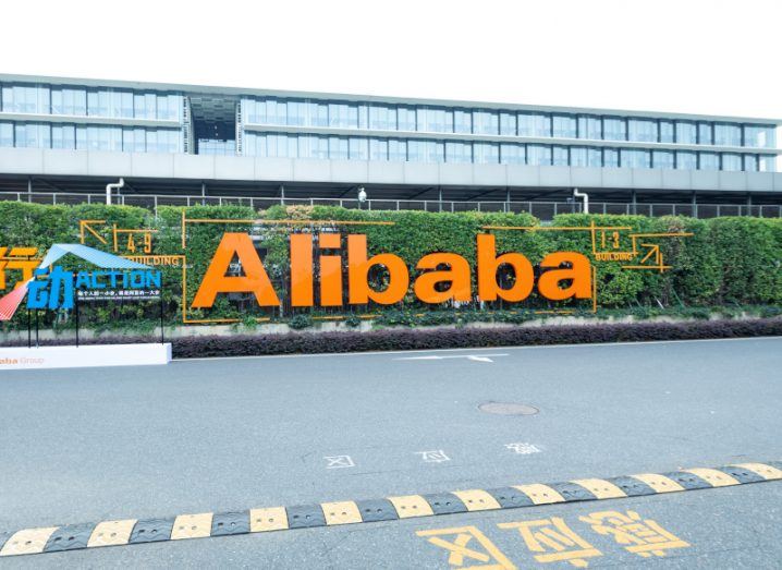 The Alibaba logo in front of a green hedge with a building and grey sky in the background.