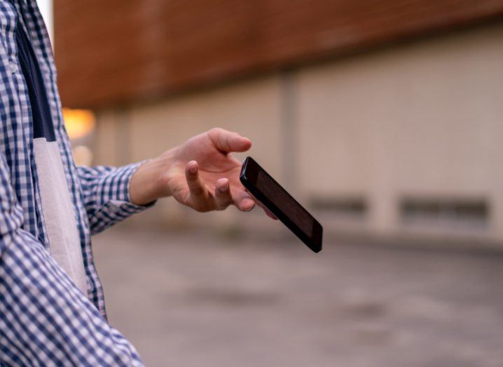 A man holding a smartphone that is slipping out of his hand, with a road and a wall in the background.