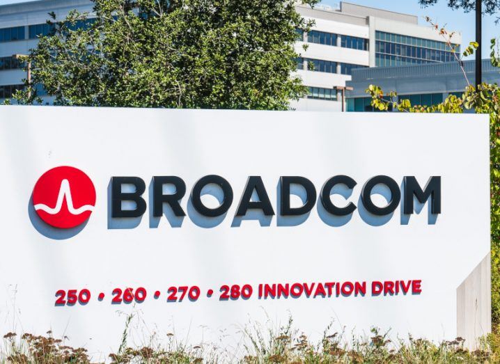 The Broadcom logo on a white sign, with small plants in front, while trees and a building are in the background.