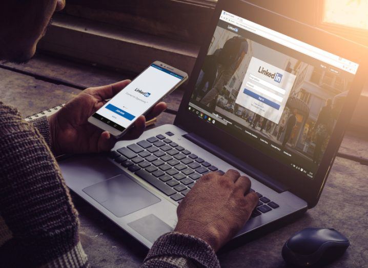 A person typing on a laptop that has the LinkedIn website on the screen. The person is holding a smartphone in his left hand that also has a LinkedIn logo on the screen.