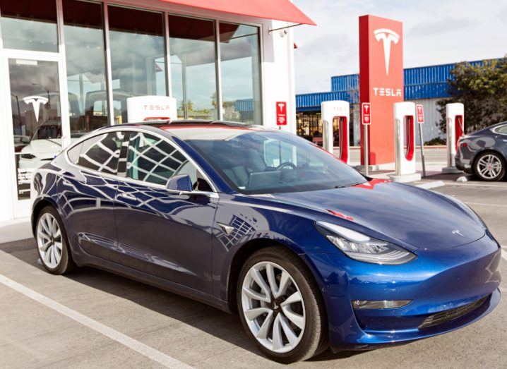 A Tesla vehicle in front of a building and charge station, with the Tesla logo on a sign in the background.