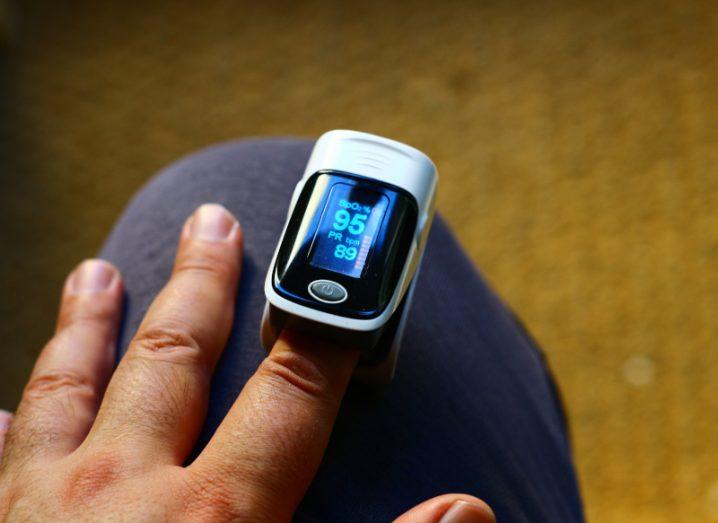 A medical device on a person's finger, reading their pulse. The person's hand is resting on their knee, with a brown floor in the background.