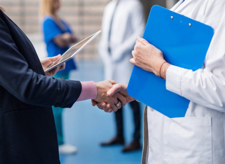 Two people shaking hands, with two other people in the background. One of the people is wearing a business suit while the other is in a white lab coat.