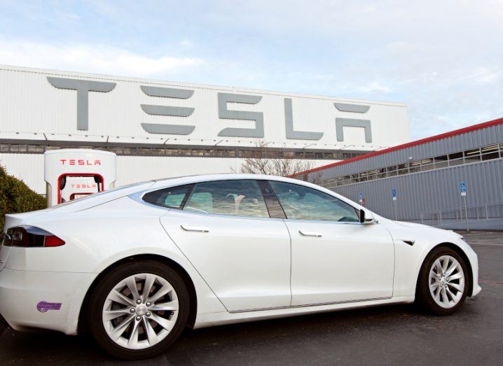 A white Tesla car in front of a large building with the Tesla logo on the side.