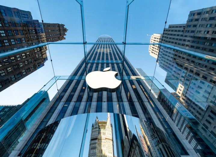 The Apple logo on the side of a tall building. The perspective of the image looks up to the blue sky, with windows and the logo connected to the tall building and other buildings nearby.
