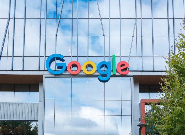 The Google logo on the front of a windowed building, with a cloudy sky being reflected from the windows.