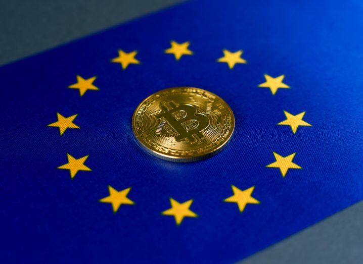 A gold bitcoin on top of an EU flag, which is laying on a grey surface.