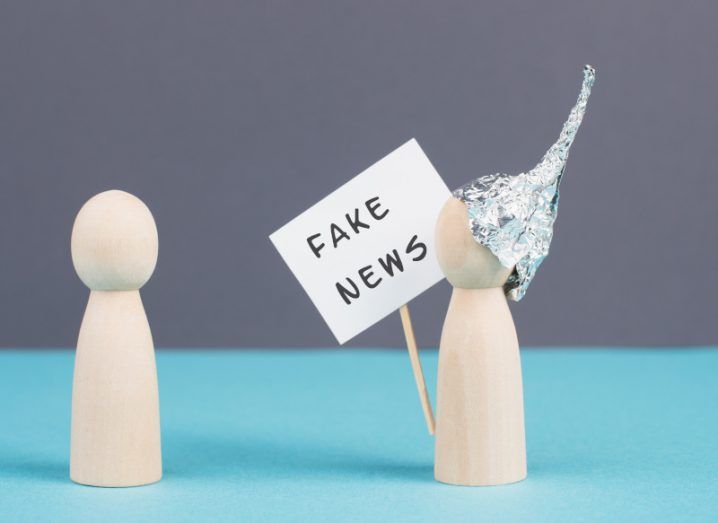 Two wooden figures, with one wearing a tinfoil hat and holding a sign that says "Fake News".