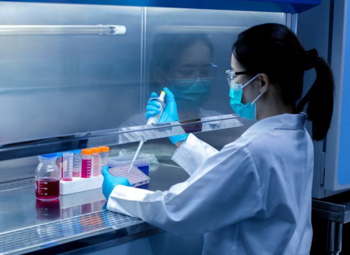 A scientist with her back to the camera working with lab equipment in a lab wearing a white coat and a medical mask.