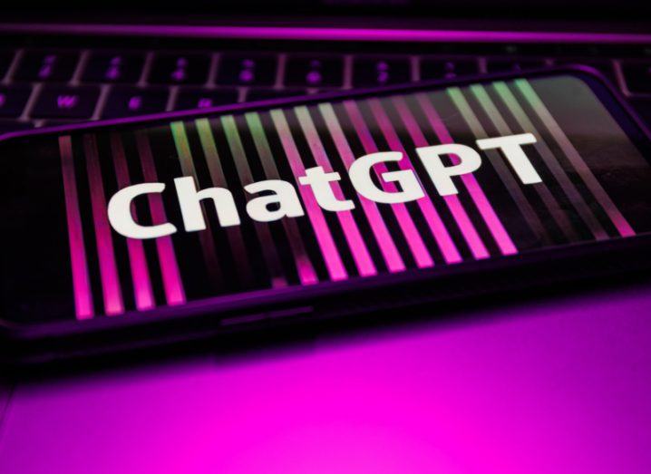 The word ChatGPT on a smartphone screen with pink lines running down the screen. The phone is on a table with pink lighting on it, with a keyboard behind it.