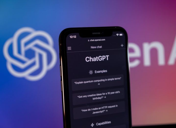 A smartphone with the ChatGPT logo on the screen and a wall with the OpenAI logo in the background.