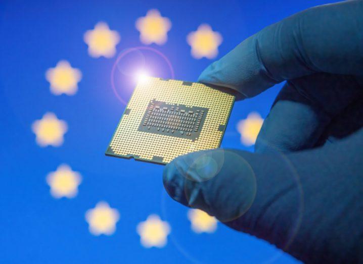 A hand in a glove holding a computer chip, with the EU flag in the background.