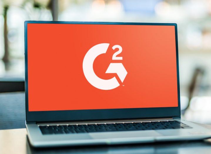 The G2 logo in an orange background on a laptop screen. The laptop is on a wooden table with a blurred background.