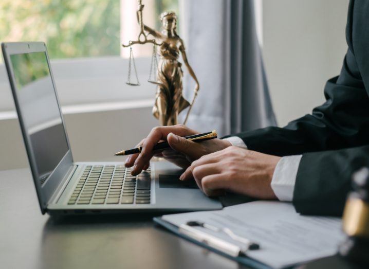 A person in a suit typing on a laptop with a statue of a woman holding scales on a table. Used to represent legal work.