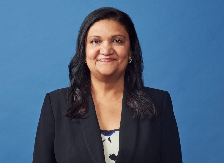 A woman with long dark hair wearing a black blazer smiles at the camera against a blue background. She is Archie Deskus, CIO of PayPal.