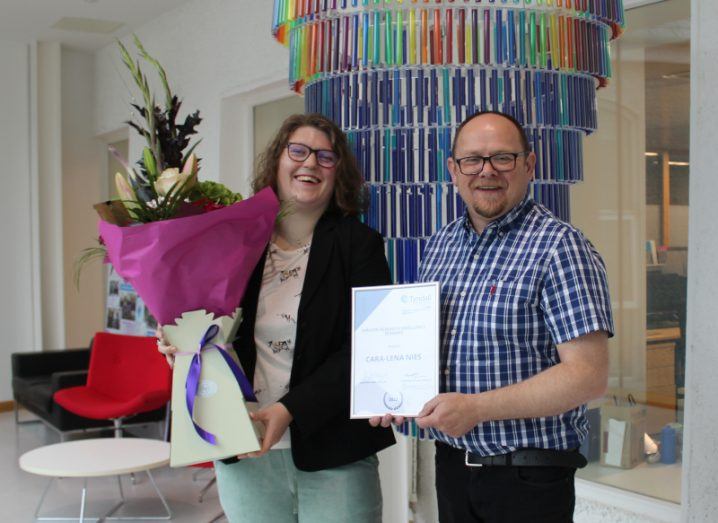 A woman with brown hair smiles, holding a bunch of flowers. A man in a blue-checked shirt also smiles. He is presenting her with a certificate.