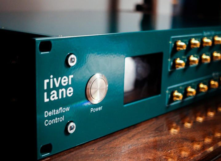 A green device with the Riverlane logo on it, called Deltaflow Control. The green device is on a wooden table.