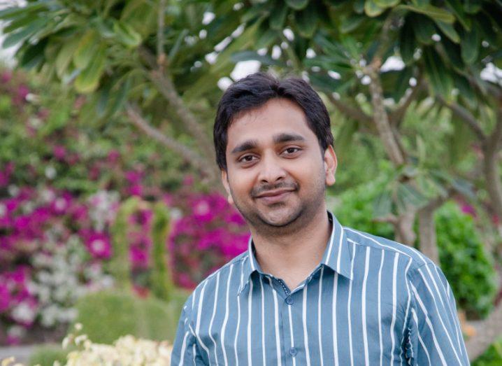 A man smiles at the camera in a blue striped shirt. There are flowers and trees behind him. We only see from his chest up.