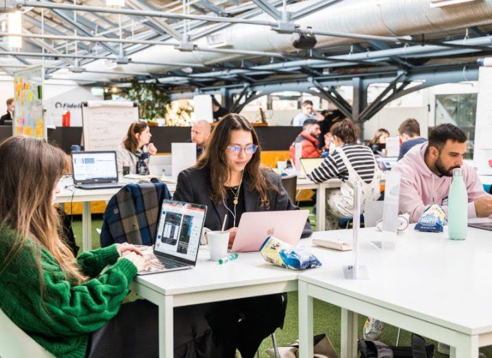 A group of people sitting in a co-working space in a brightly lit space working on laptops.