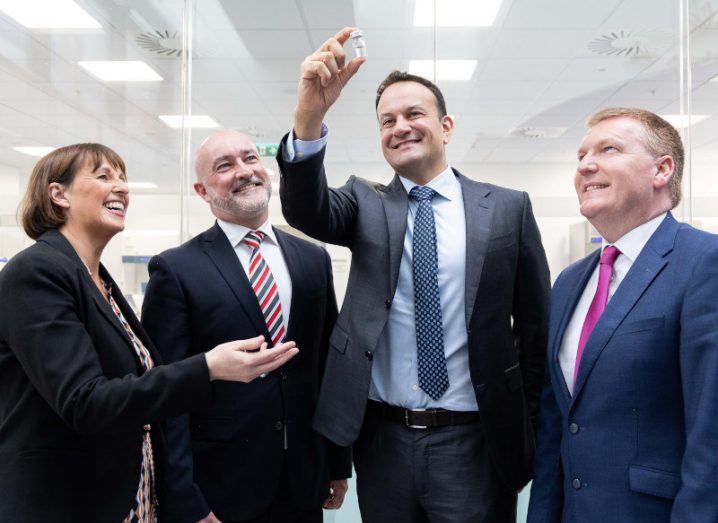 Four people with one of them holding a little medicine vial aloft. They are all standing in a white room.