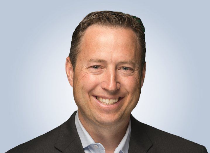 A headshot of a man in a suit smiling at the camera against a blue background. He is Telus International’s Michael Ringman.