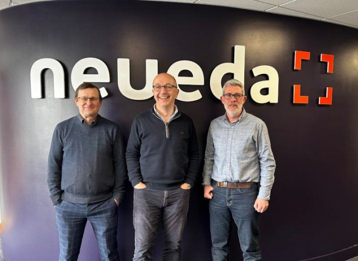 Three men standing together in front of a wall with the Neueda logo on it.