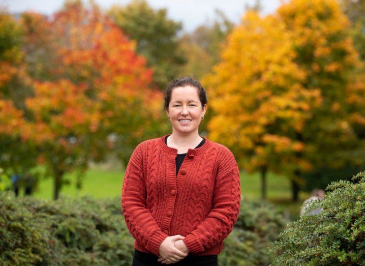 A woman stands in a red cardigan and smiles at the camera. There are autumn trees and shrubbery behind her.