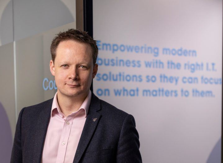 Headshot of a man wearing a suit and standing in an indoor space. Some text is visible on the wall behind him. The man is Kevin O'Loughlin, CEO of Nostra, which just acquired Passax.