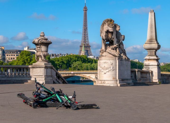 A pile of e-scooters lying on the ground in Paris with the Eiffel Tower visible in the distance.