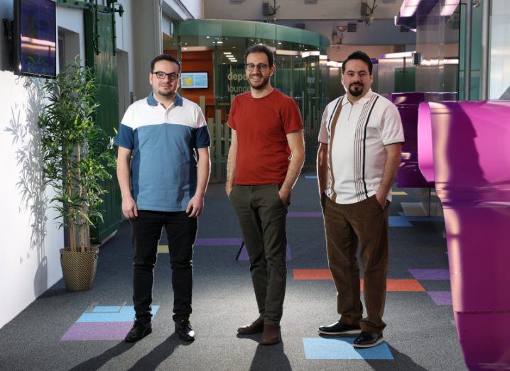 The three founders of Perfogram stand next to each other in a corporate-looking office space with plants and TV screens in the background.