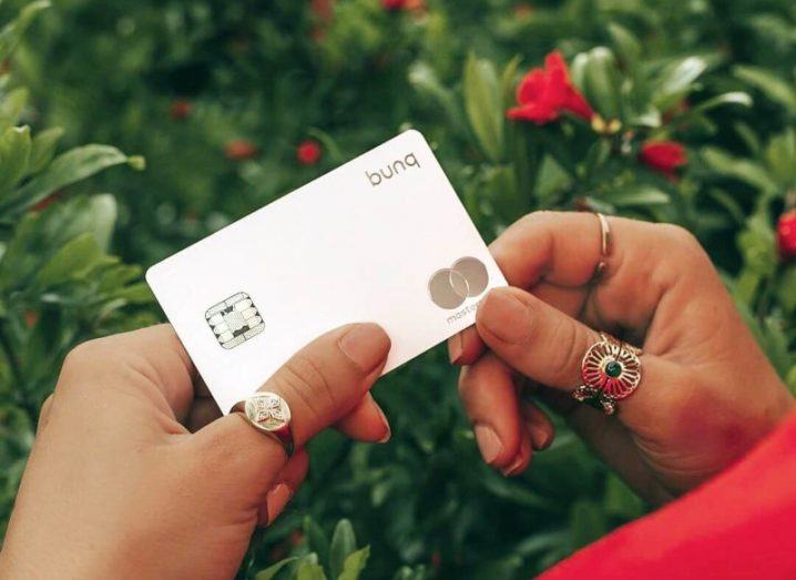 A person's hands holding a grey metallic credit card with the Bunq logo on the top right. There are plants in the background.